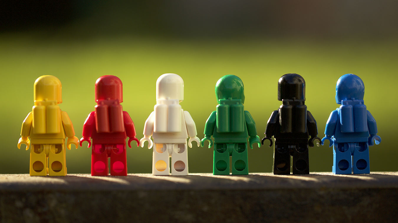Six classic LEGO spacemen are lined up in a row facing away from the viewer. All six colors of the spacemen are represented in this outdoor toy photograph.