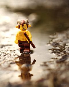 A Lego faun walks through a puddle holding his flute. His reflection mingles in the water and the background bokeh.