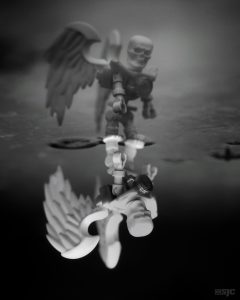 A lego skeleton with wings stands in a puddle and his ghastly reflection stares back out to the viewer in this black and white outdoor toy photograph.