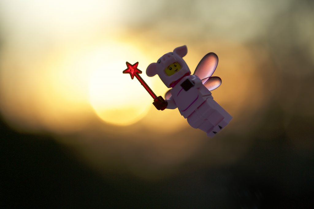 A LEGO pig mini figure with pink wings, holding a ed wand flies across the rising sun in this short depth of field outdoor toy photograph.