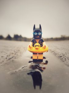 lego batman, dressed in swim wear, stands in a puddle looking for the beach