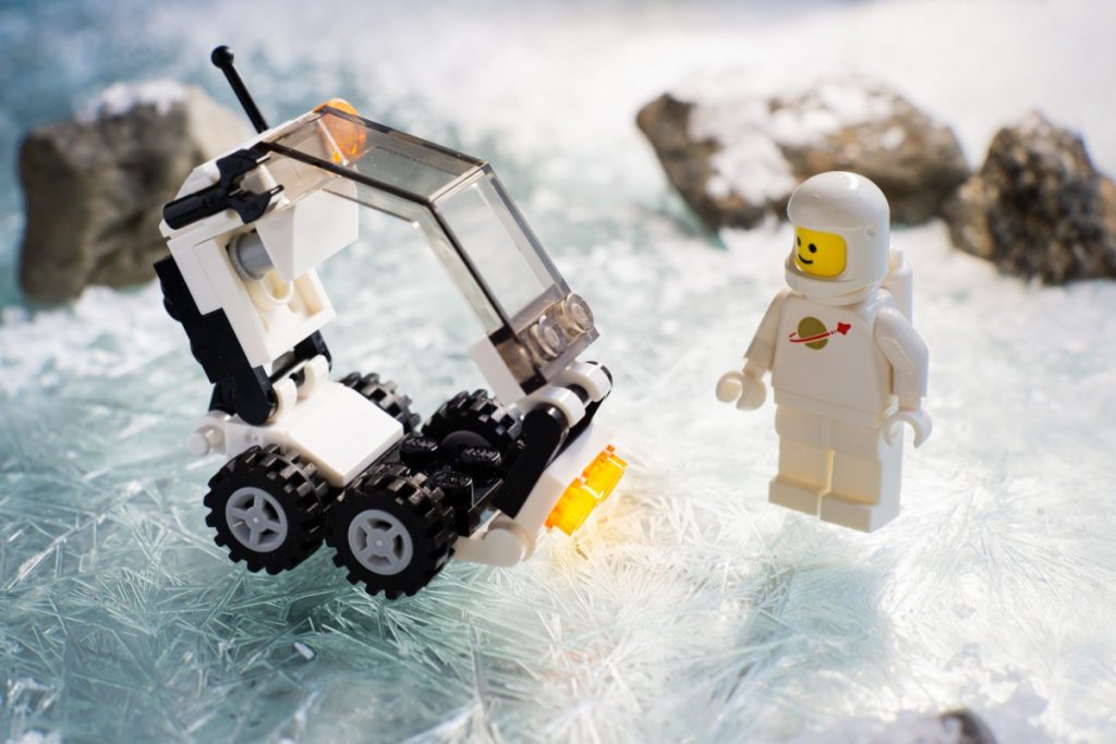 white classic LEGO spaceman stands on a ice surface next two a small brick built transport vehicle. by Anna Bitanga