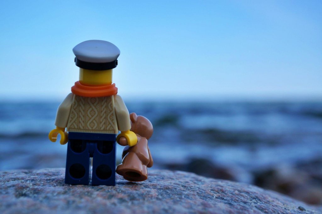 A Lego captain minifigure, shot from behind, is holding a Lego Teddy Bear and is looking on the sea