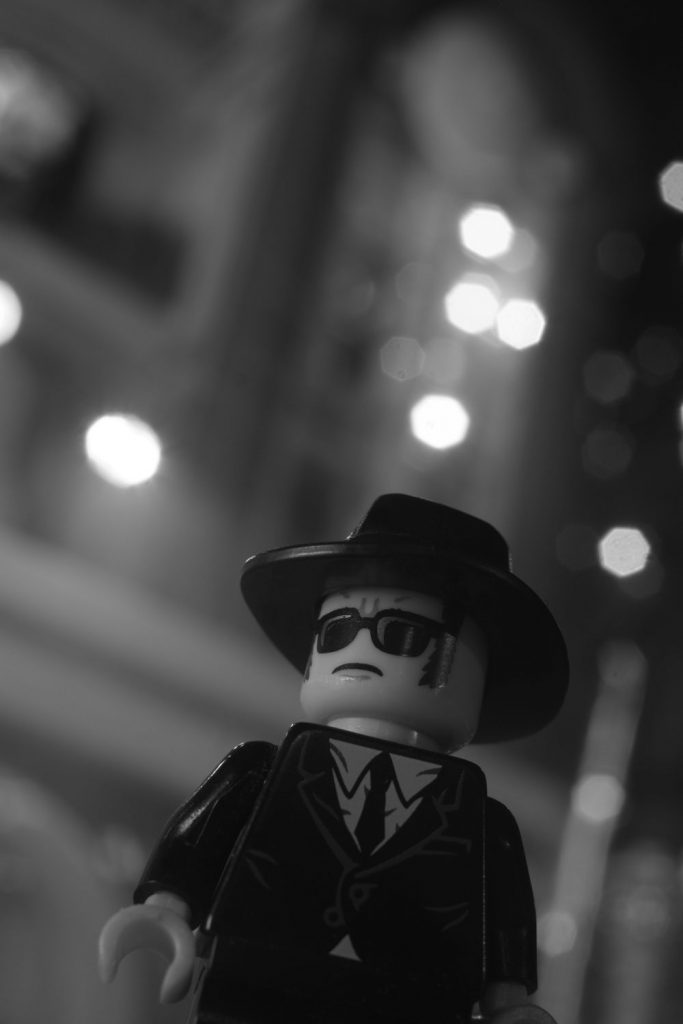 Lego minifigure in sunglasses, black suit and fedora hat in noir-like dutch-angle shot