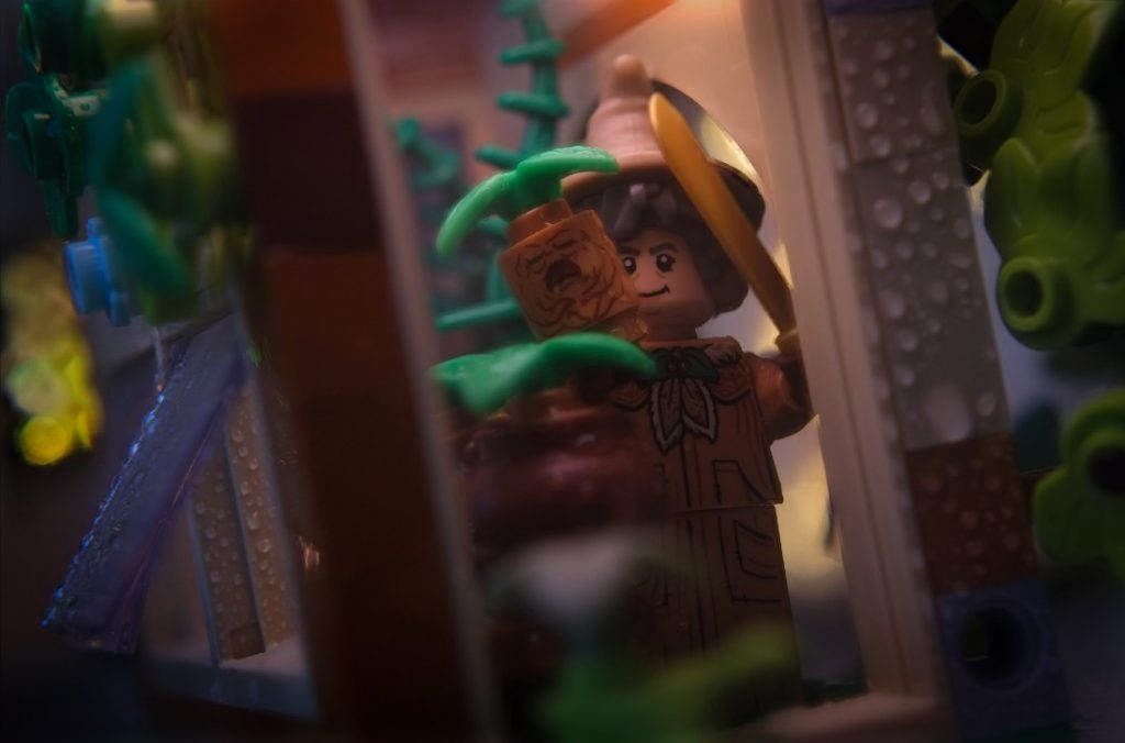 Pomona Sprout LEGO minifigure standing inside Greenhouse, holding a mandrake in right hand and machete in left hand.