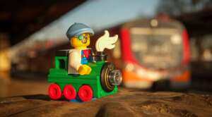 A LEGO minifigure of a boy in the locomotive costume, posing with train in the background, on the train station