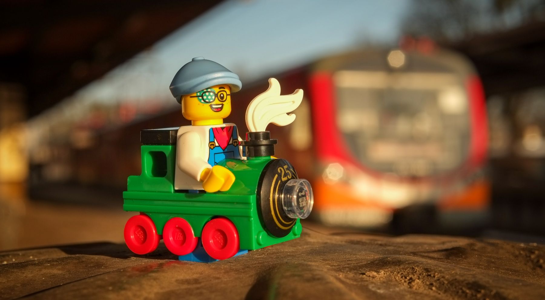 A LEGO minifigure of a boy in the locomotive costume, posing with train in the background, on the train station