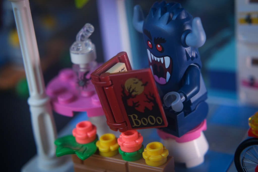 Bogeyman minifigure is sitting at a table in front of a cafe, reading a book with the word BOOO written on it and a crooked trees and a full moon on the cover.