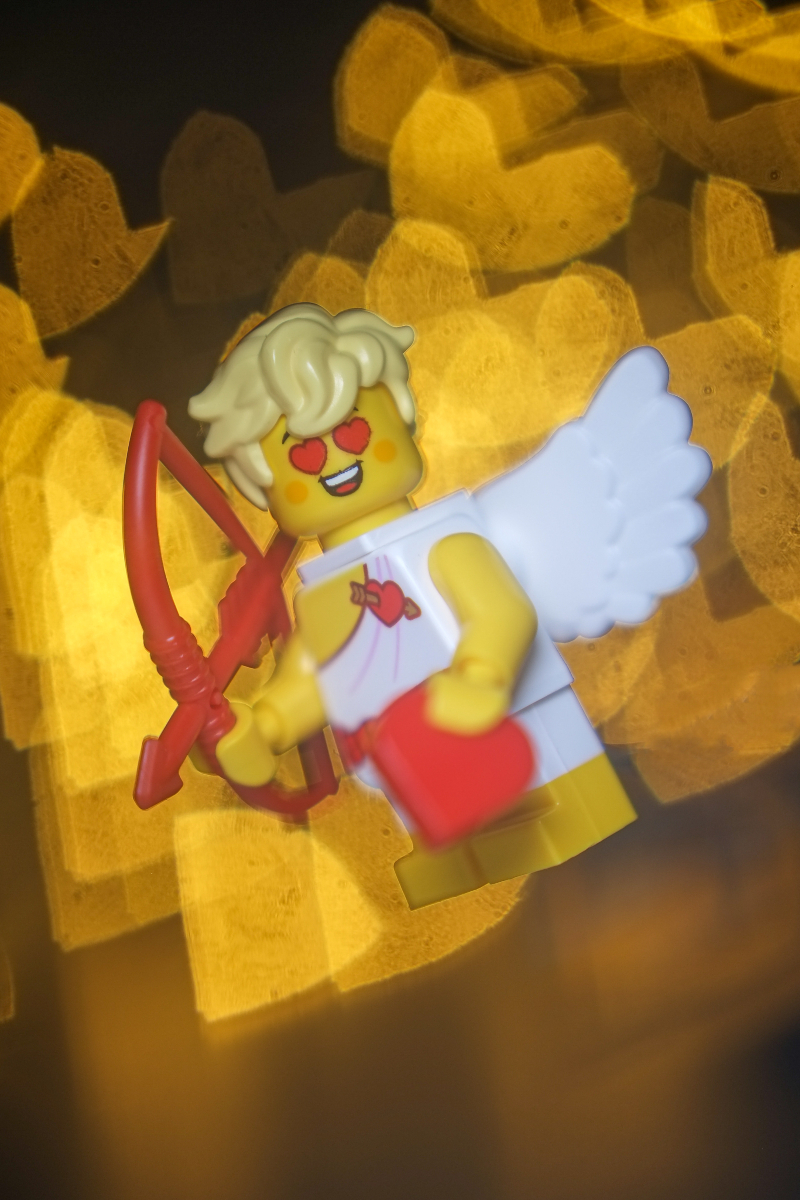 A laughing Cupid minifigure with wide open heart-shaped eyes flies against an intense heart-shaped bokeh background.